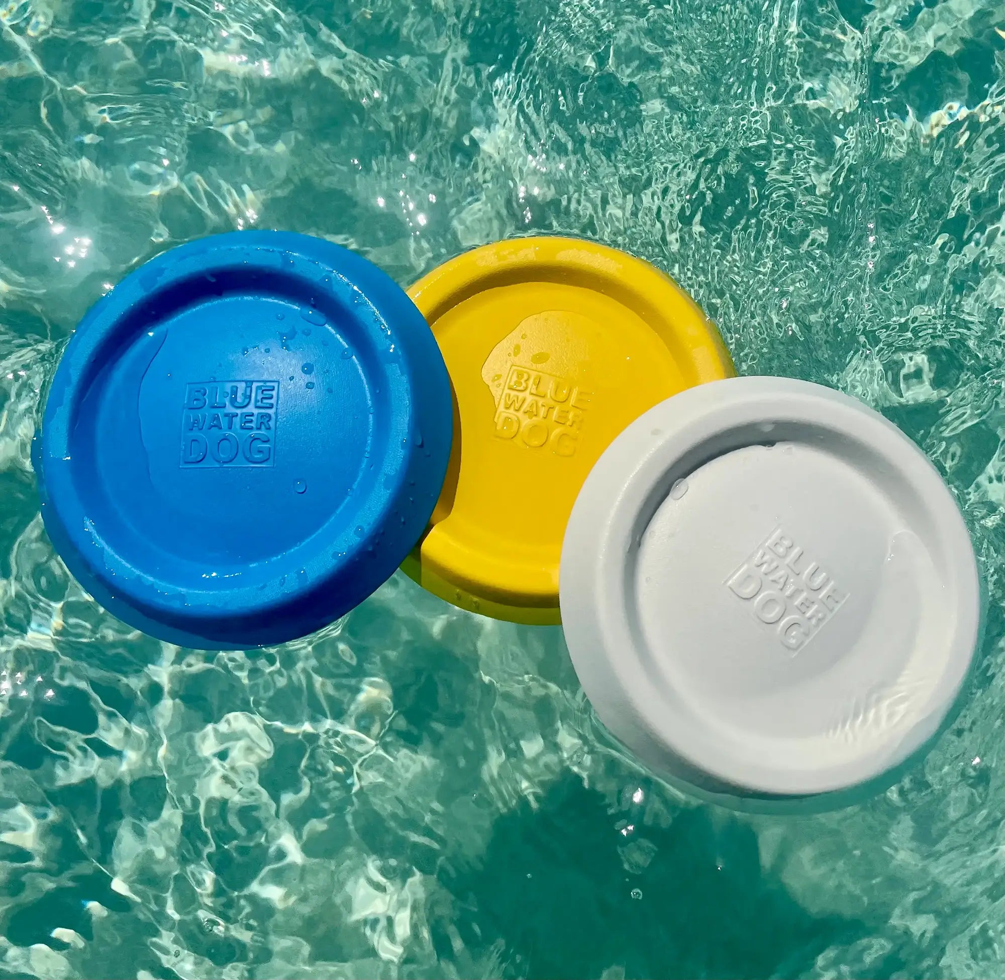 Three dog frisbees in the colors of blue, yellow, and white floating together in light blue ocean water.